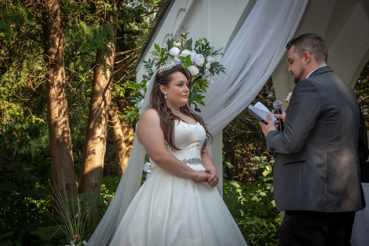 A bride in white stands under a floral arch as an officiant reads from a booklet, sunlight filtering through trees.
