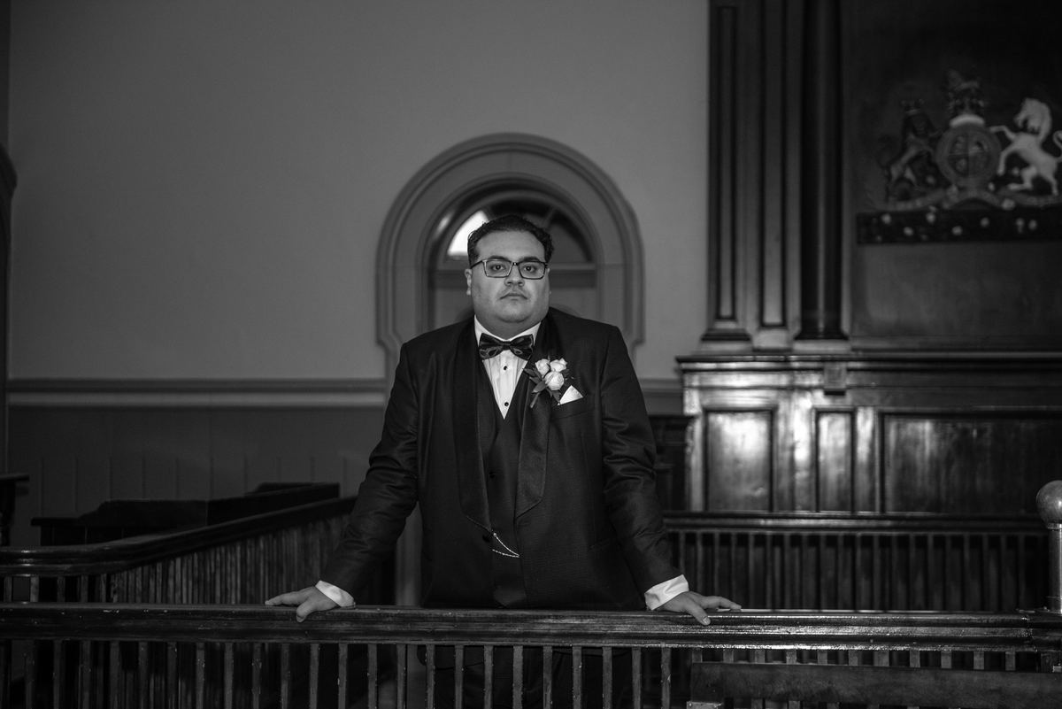 Groom in formal attire stands at church pew railing, looking directly at camera during wedding ceremony.