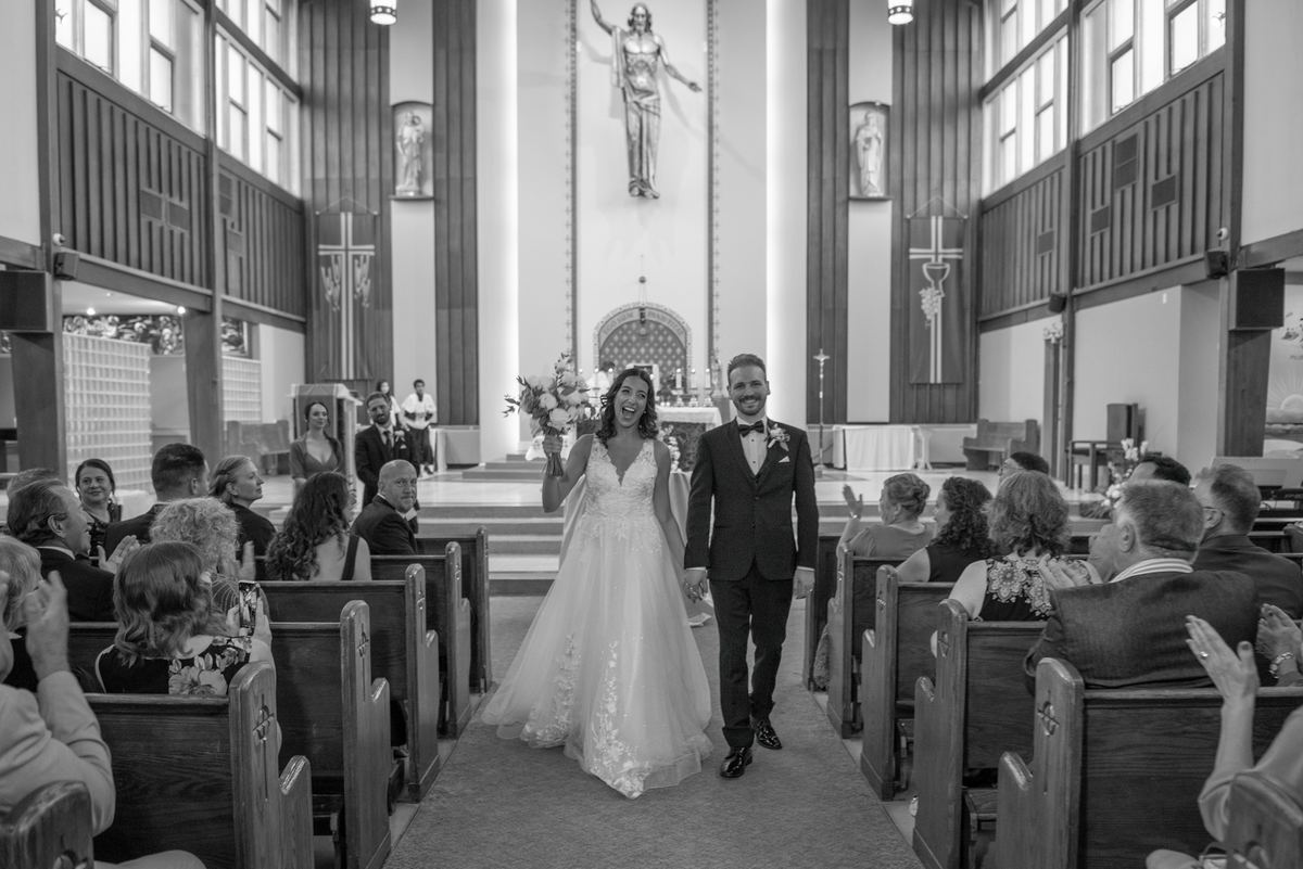 A smiling bride and groom exit a church, holding hands and waving to applauding guests as soft light filters through windows.