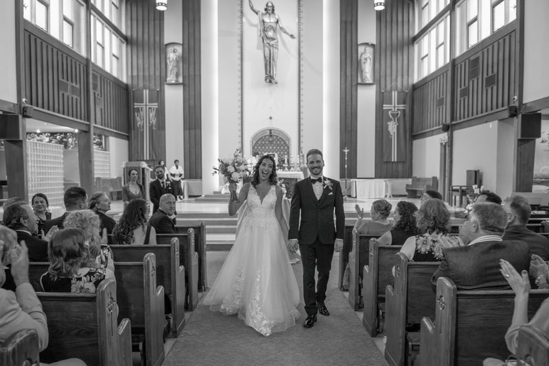 A smiling bride and groom exit a church, holding hands and waving to applauding guests as soft light filters through windows.