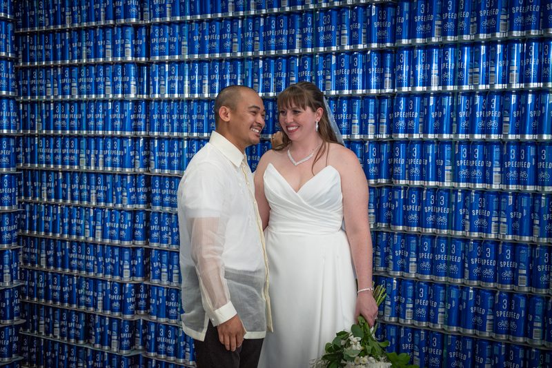 Samantha and Robbie smile joyfully, standing close together in front of a towering wall of blue 'Speedee' soda cans.