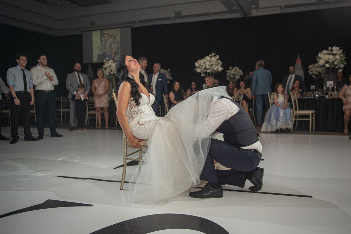 Bride and groom dance joyfully, with the groom lifting her as she leans back, guests clapping in background.
