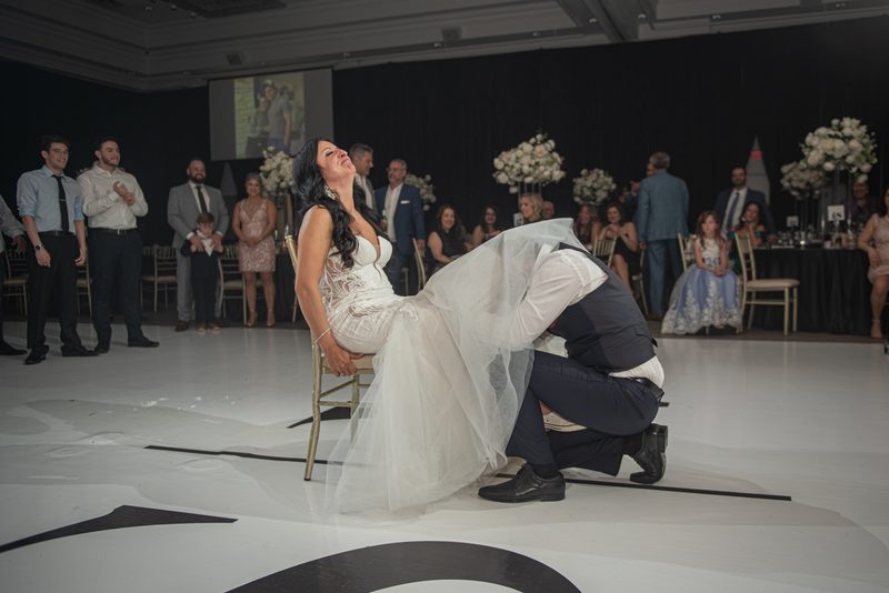 Bride and groom dance joyfully, with the groom lifting her as she leans back, guests clapping in background.