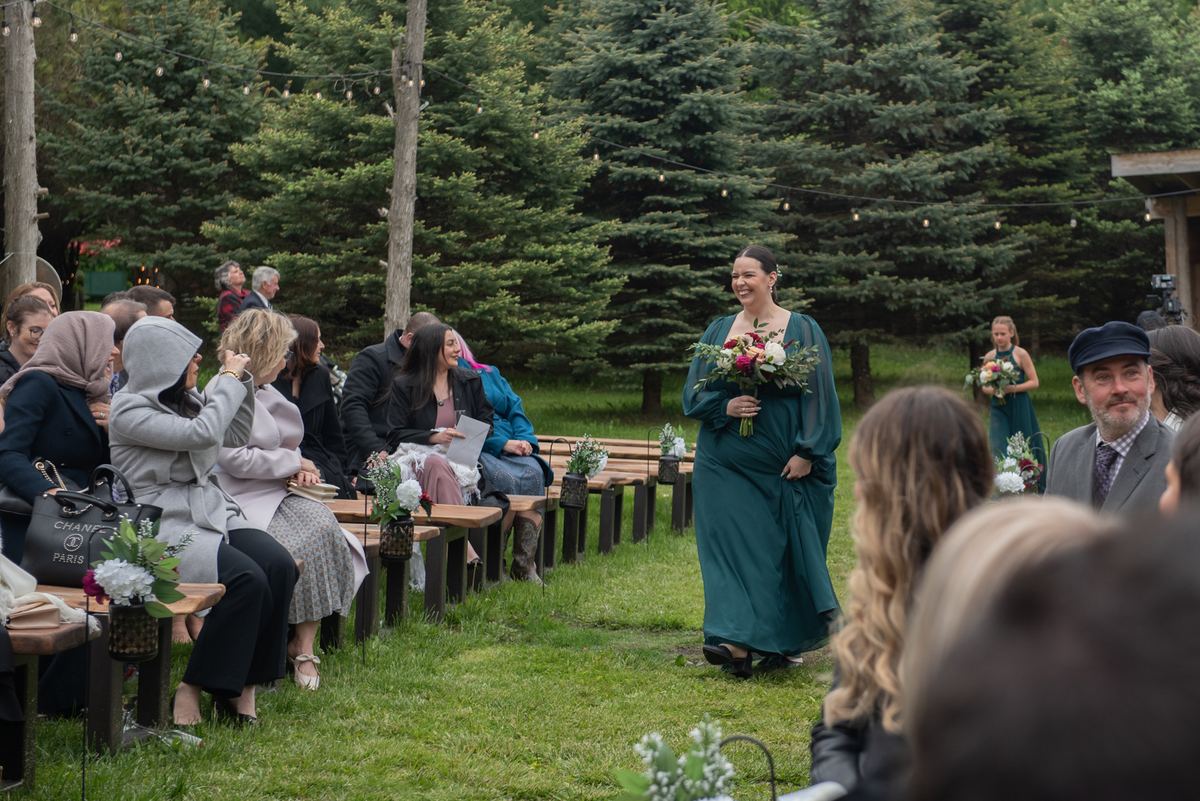 A smiling bridesmaid in teal walks down the grassy aisle toward seated guests under evergreen trees.