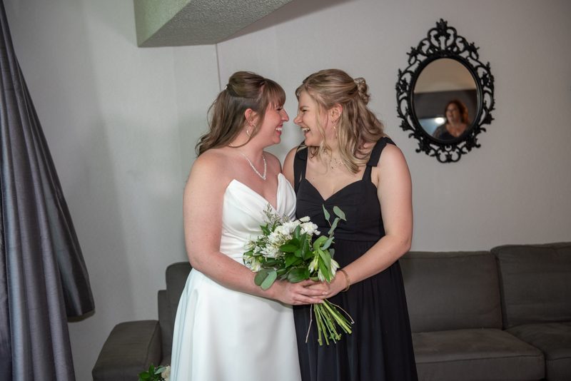 Two brides laughing and embracing while holding a bouquet in an indoor living space with a sofa and curtain.