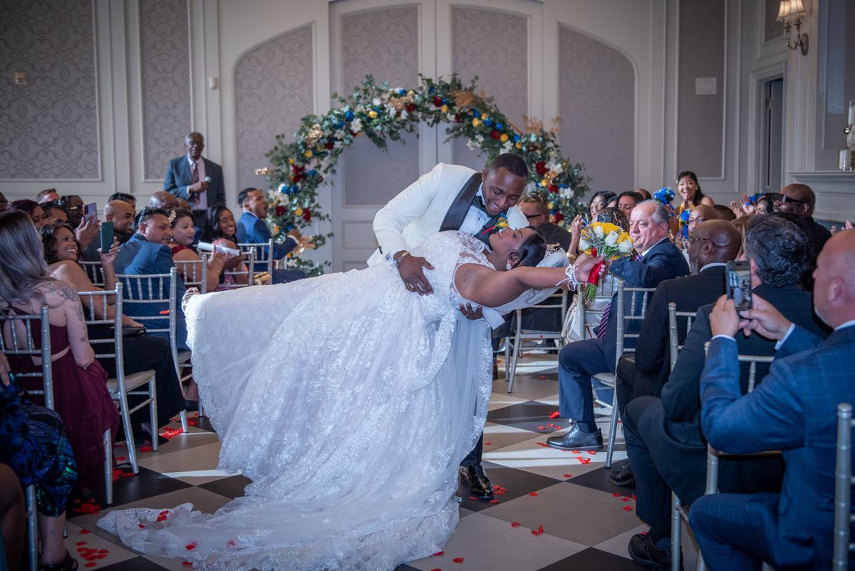 The groom lifts the bride as they dance down the aisle, surrounded by guests and red petals, under a floral arch.