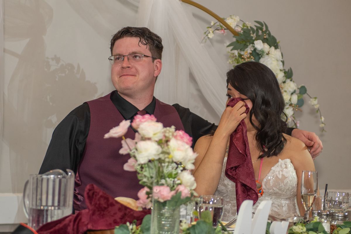 Bride wiping tears with burgundy cloth, groom smiling beside her with arm around her, in warmly lit reception space.