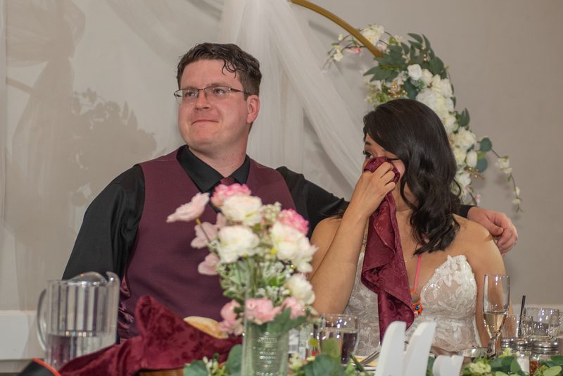 Bride wiping tears with burgundy cloth, groom smiling beside her with arm around her, in warmly lit reception space.