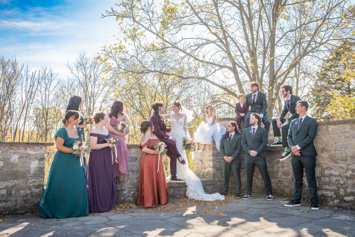 A bride in white and her wedding party gather outdoors beside a stone wall under golden autumn light, smiling.