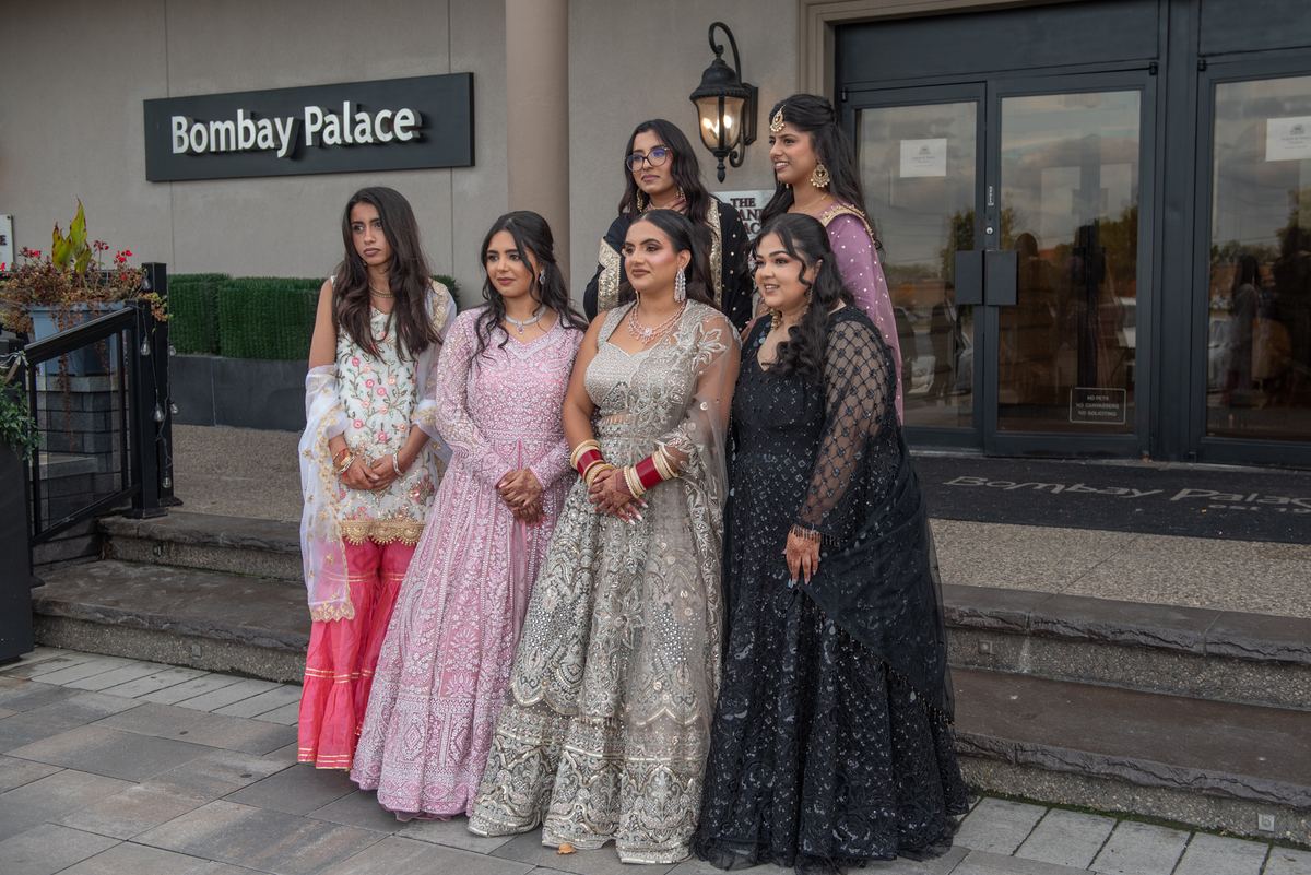 Six elegantly dressed South Asian women pose on steps outside a modern building, glass doors visible behind them.