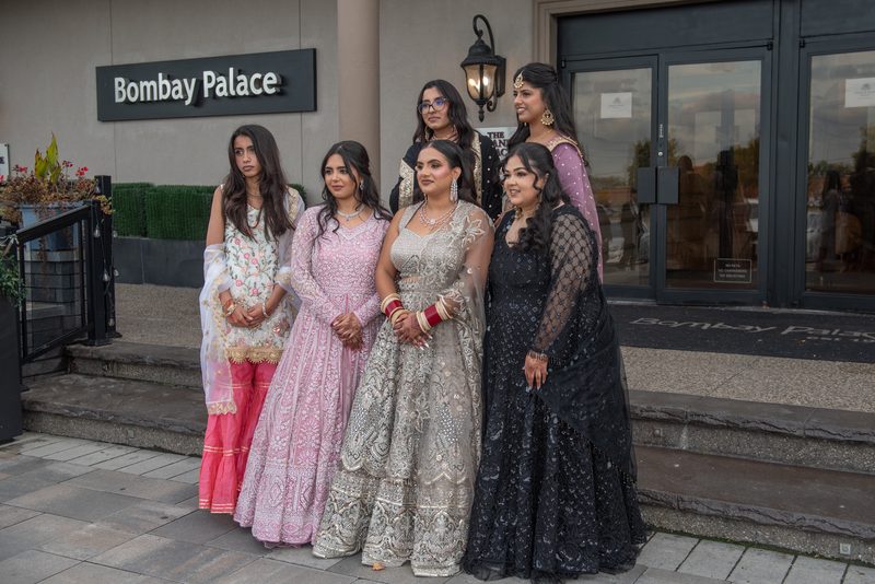 Six elegantly dressed South Asian women pose on steps outside a modern building, glass doors visible behind them.