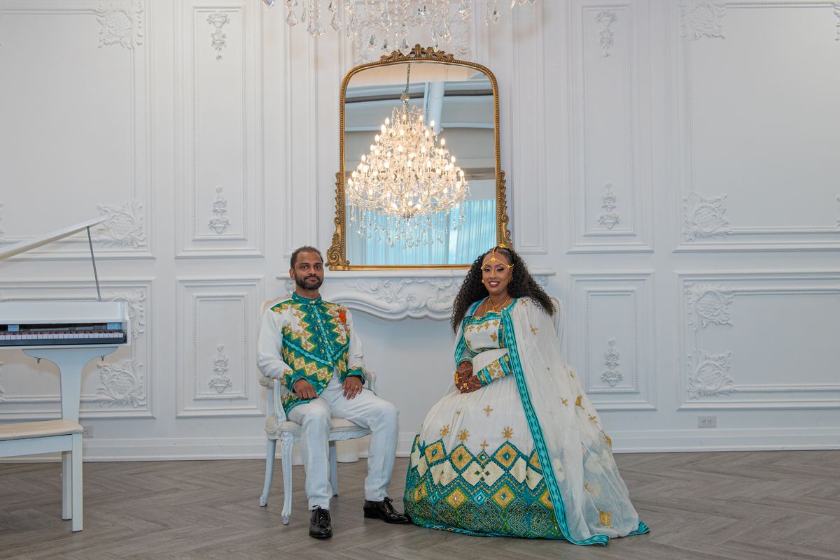 A couple in traditional attire sit for a portrait in an ornate room with a grand chandelier and gilt-framed mirror.