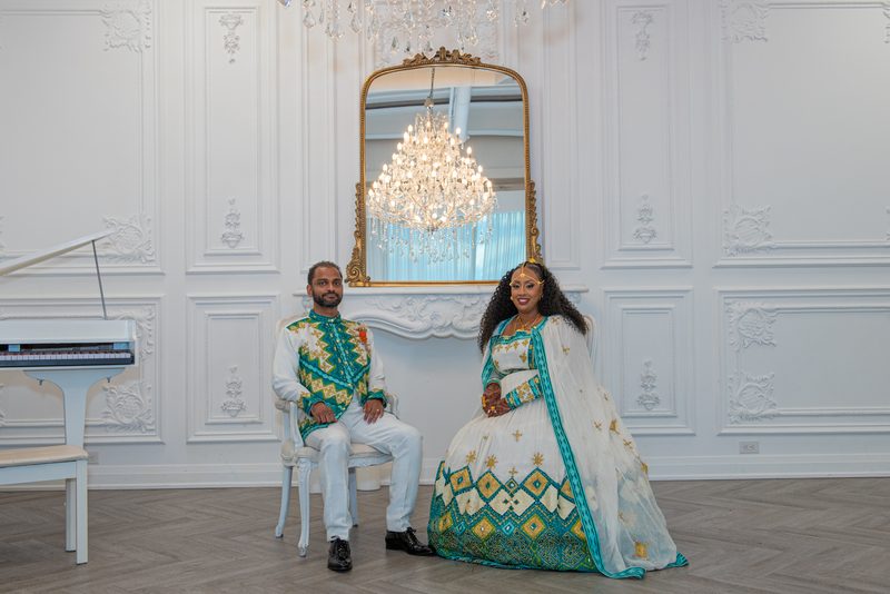 A couple in traditional attire sit for a portrait in an ornate room with a grand chandelier and gilt-framed mirror.