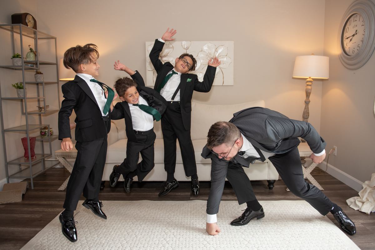 A groom and three young boys in formal black suits with green ties pose energetically in a warmly lit living room.