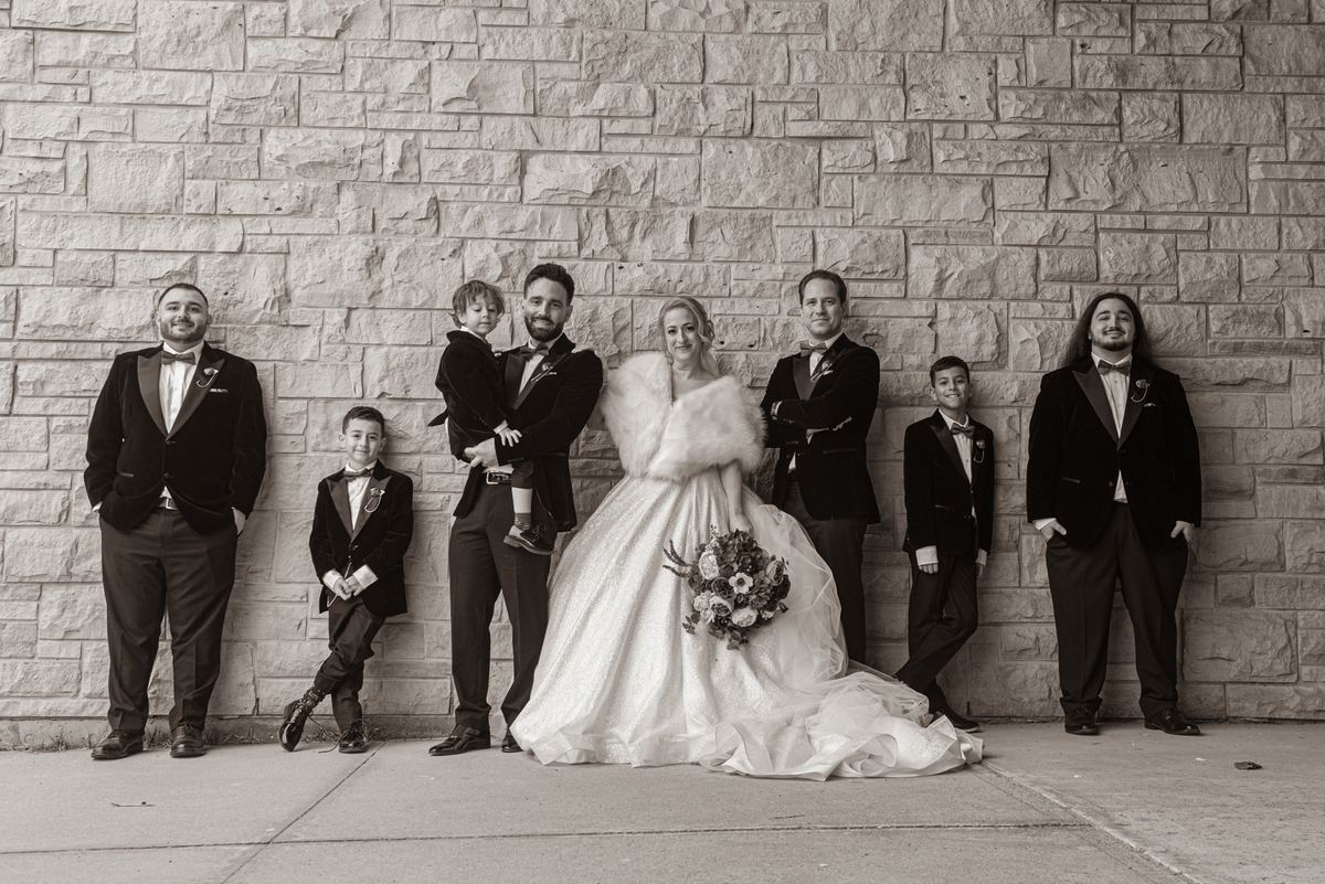 A bride in a voluminous gown and a group of formally dressed men and boys pose against a textured stone wall.