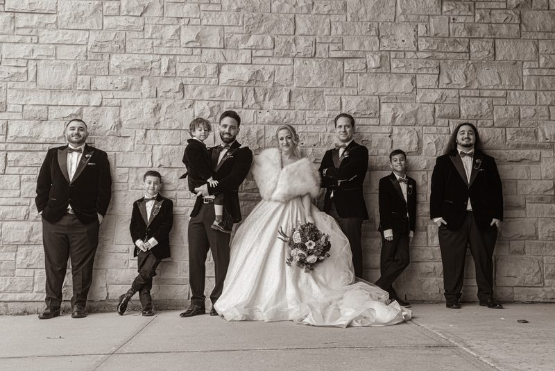 A bride in a voluminous gown and a group of formally dressed men and boys pose against a textured stone wall.