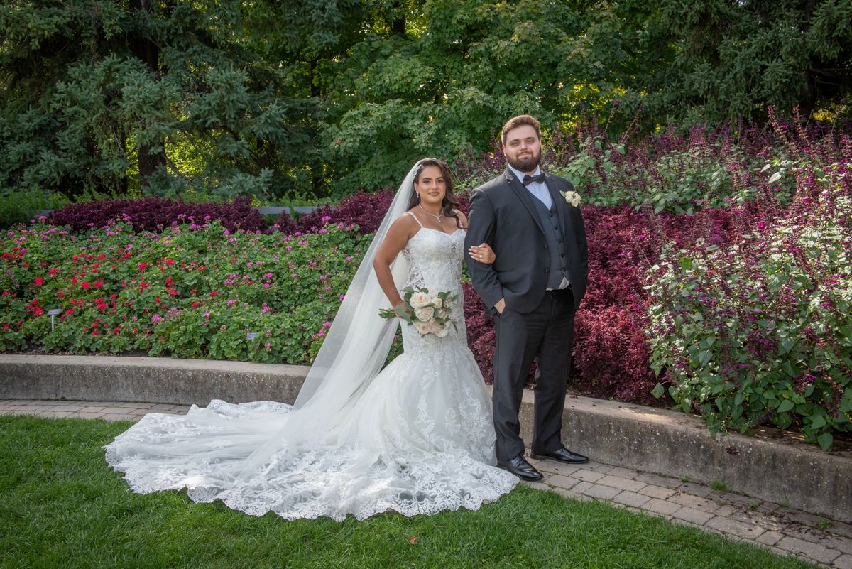 A bride in a lace gown and veil stands with her groom in a dark suit, both posing in a garden filled with vibrant flowers.