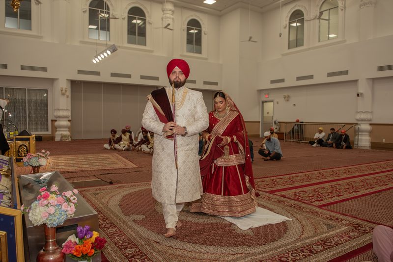 A Sikh groom and bride walk barefoot on a carpet inside a gurdwara, surrounded by guests and floral arrangements.