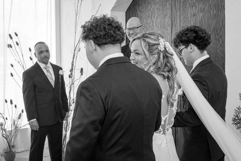 Seen from behind, a bride in a long veil walks toward her groom during the ceremony as guests watch, in black and white.