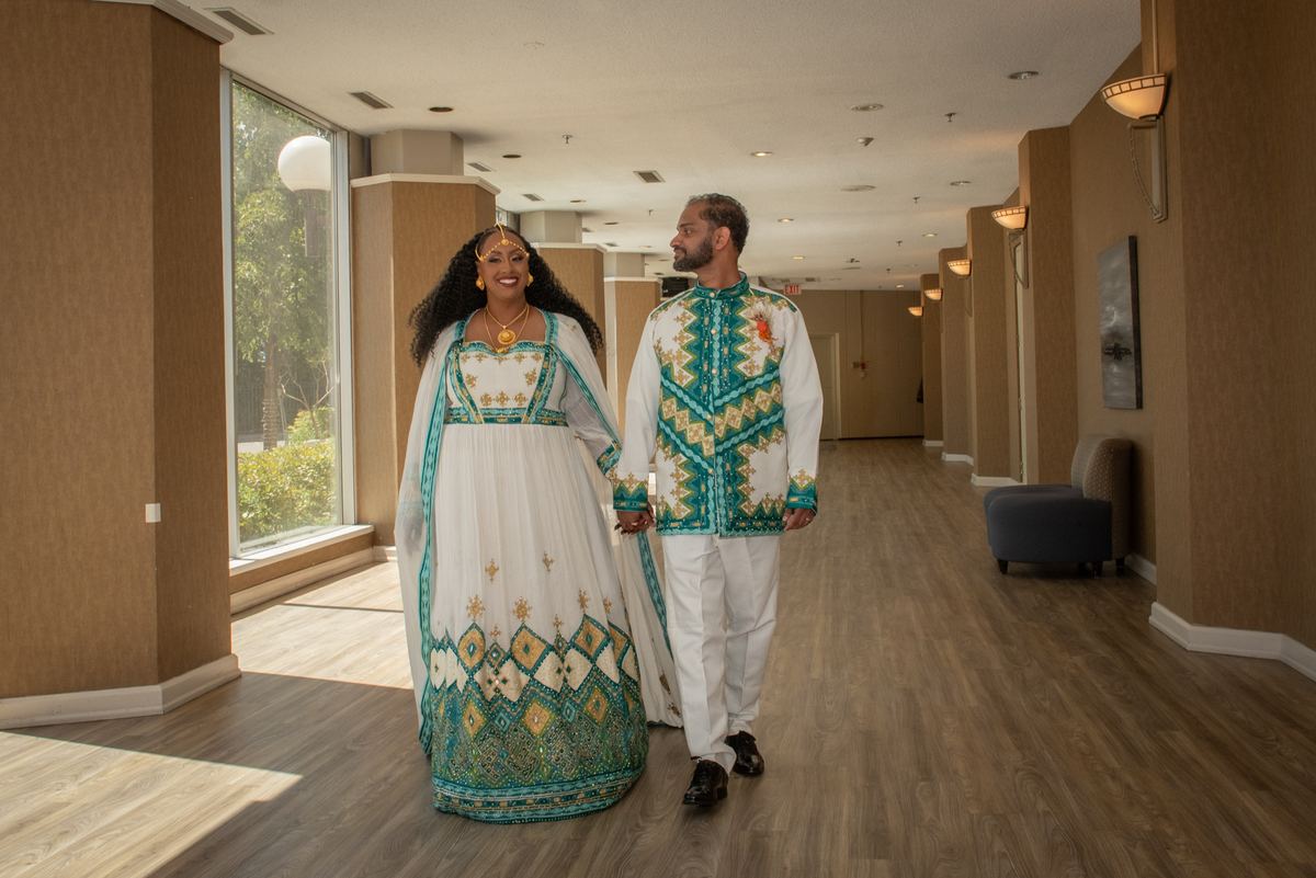 A smiling couple in traditional attire walks down a lit hotel corridor, natural light streaming through a window.