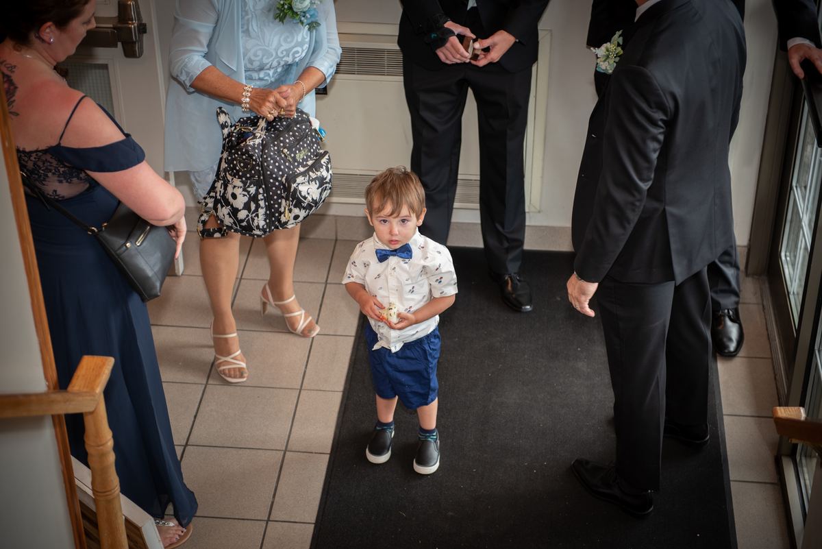 A young boy in formal attire stands on a black runner, holding a small piece of cake as adults observe.