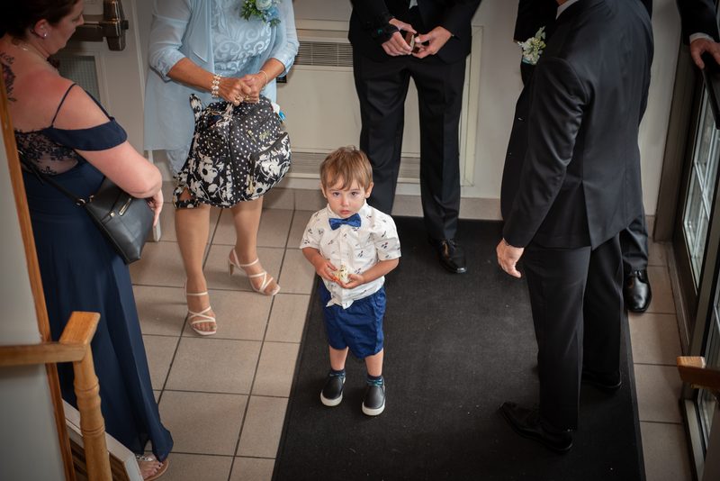 A young boy in formal attire stands on a black runner, holding a small piece of cake as adults observe.