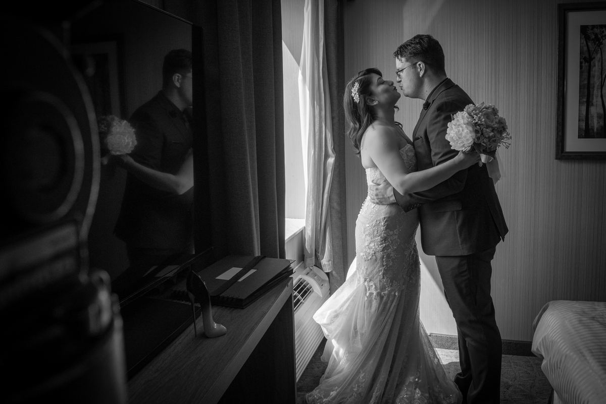 A bride and groom embrace near a window in a hotel room, illuminated by soft natural light streaming in from the right.