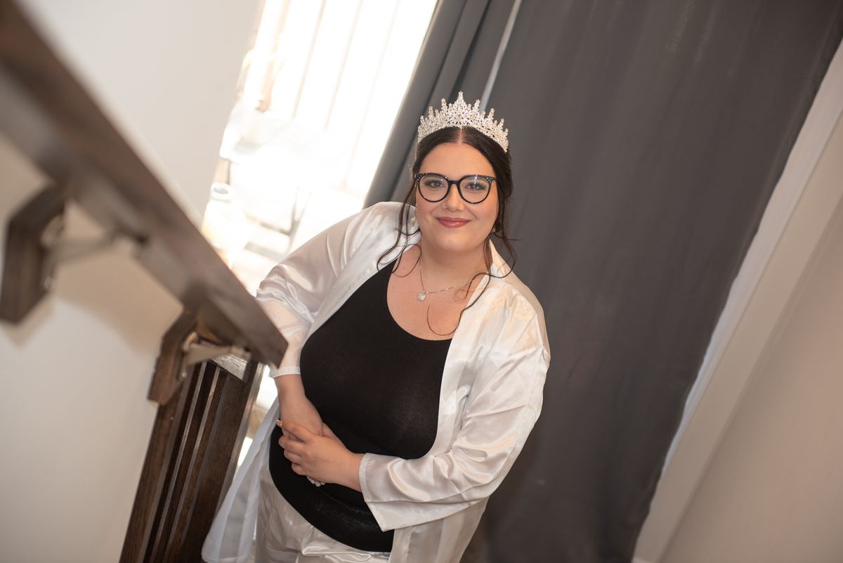 A bride smiles by a staircase, wearing a tiara and white satin robe over a black top, illuminated by soft natural light.