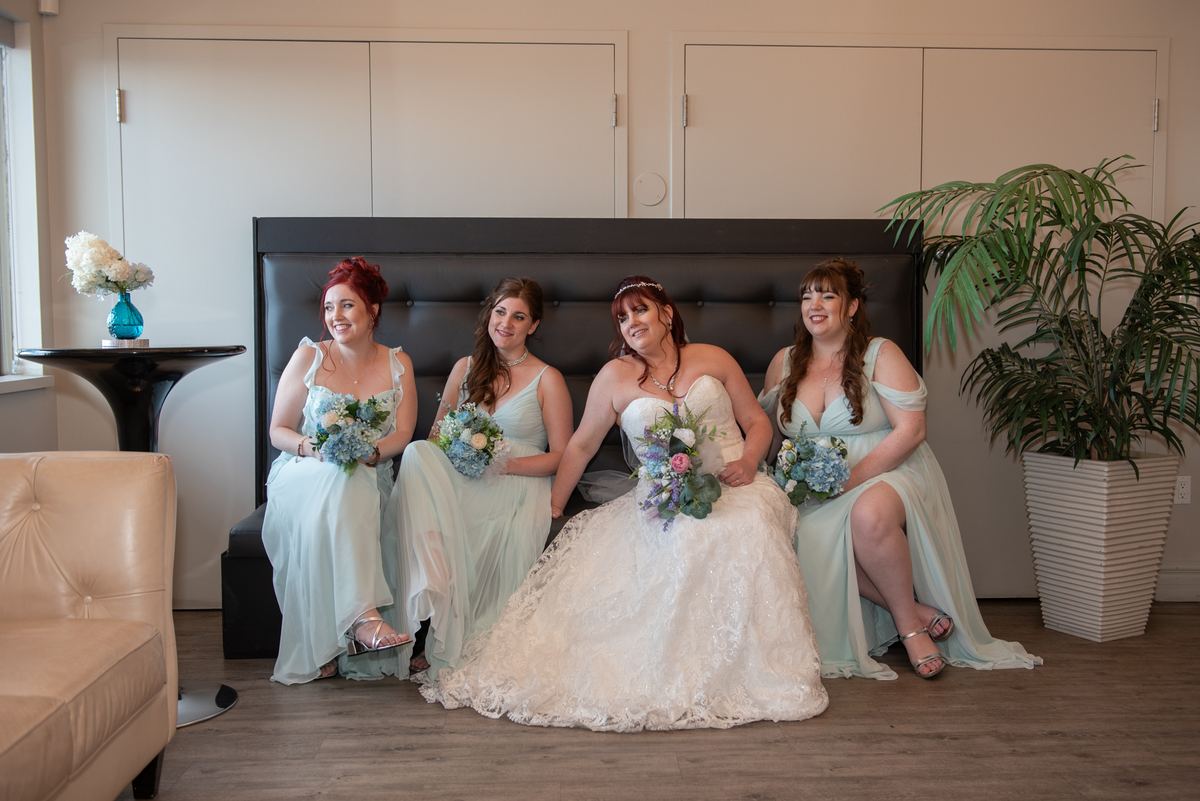 Four women, including a bride in a white gown, sit on a black bench holding flowers near a potted plant and table.