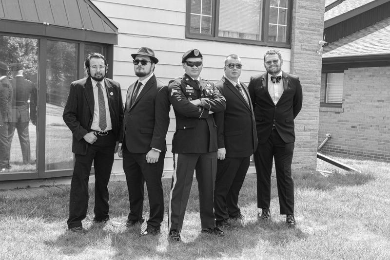 Five formally dressed men, including a soldier in uniform, stand outdoors on grass in front of a building during the day.