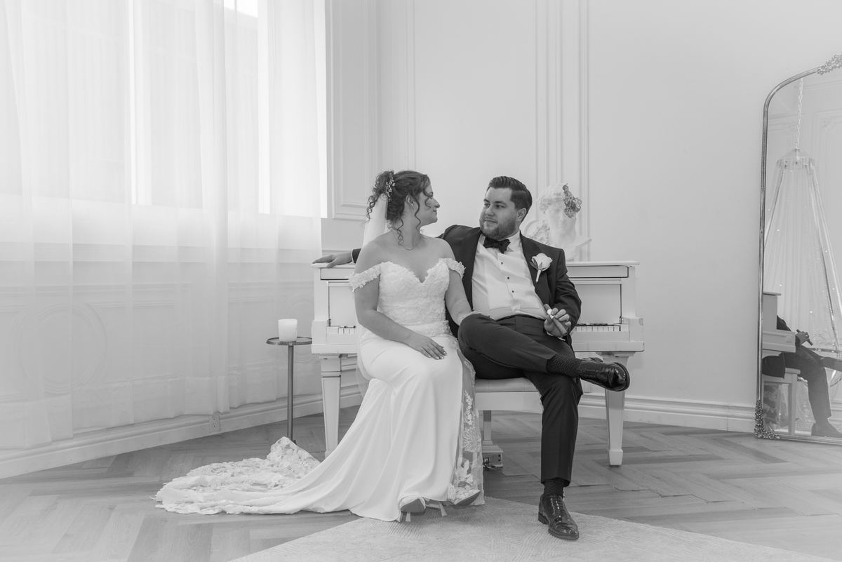 A newlywed couple sits beside a white grand piano, gazing lovingly at each other in an elegant, softly lit room.