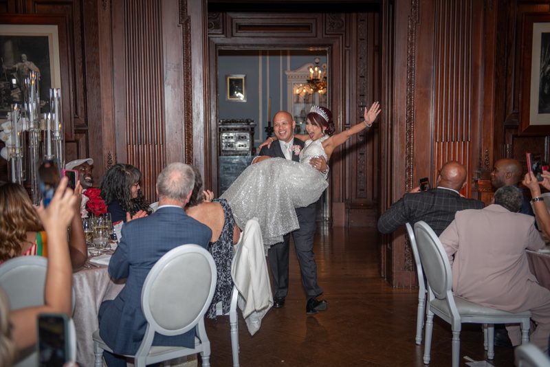 In a grand wood-paneled hall, the groom lifts his bride as she laughs and waves to guests at tables.