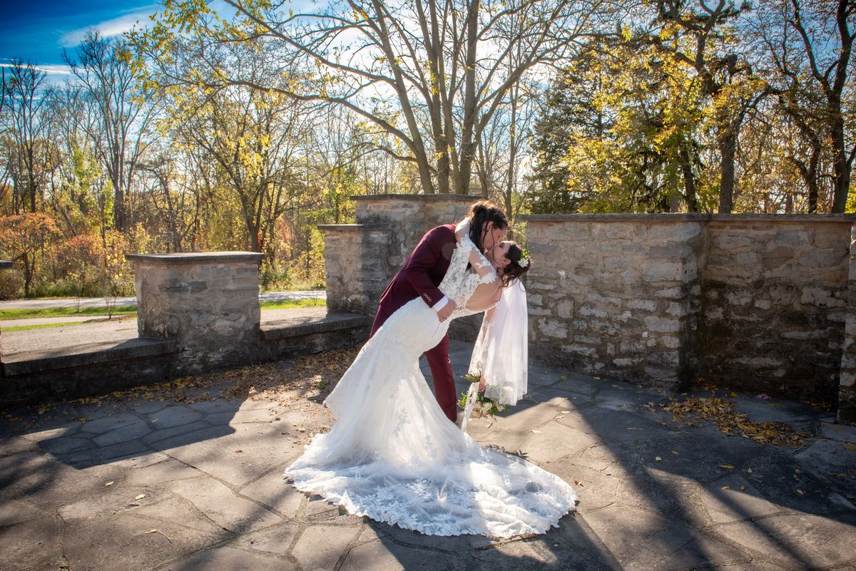 A bride and groom kiss joyfully in an autumn courtyard surrounded by trees and a stone wall, bathed in golden light.