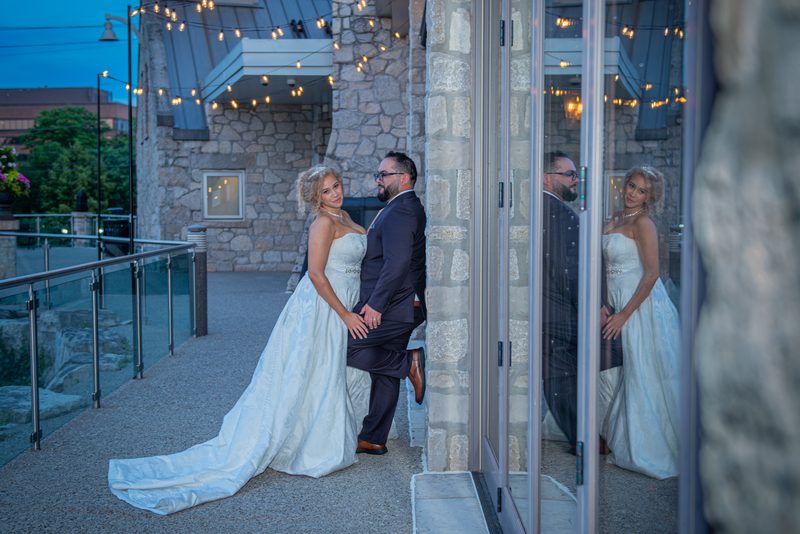 Bride and groom peek around stone pillar, their reflection visible in glass door, under string lights at dusk.