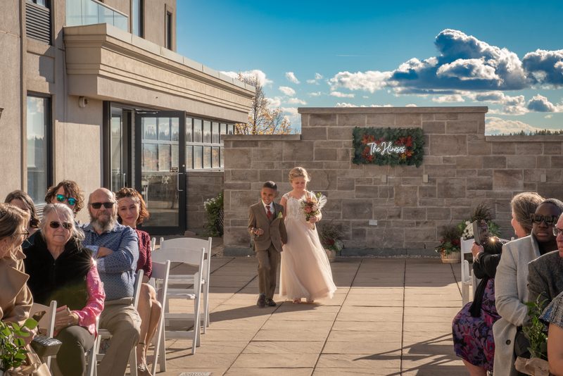 A flower girl and a boy in suits walk down an aisle toward a ceremony, flanked by guests under a bright sky.