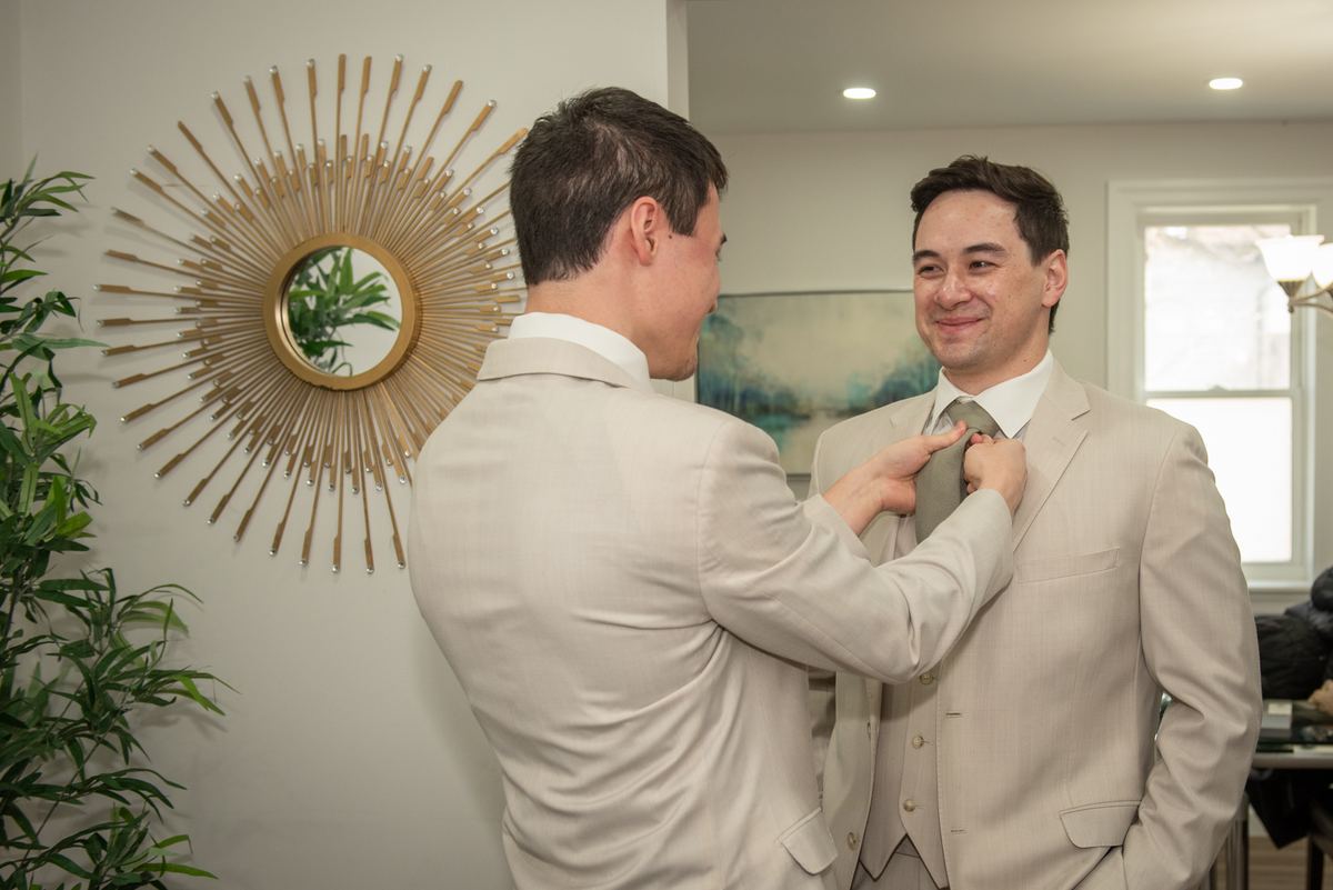 Two men in light-colored suits share an affectionate moment as one adjusts the other's tie, enhancing their bond.