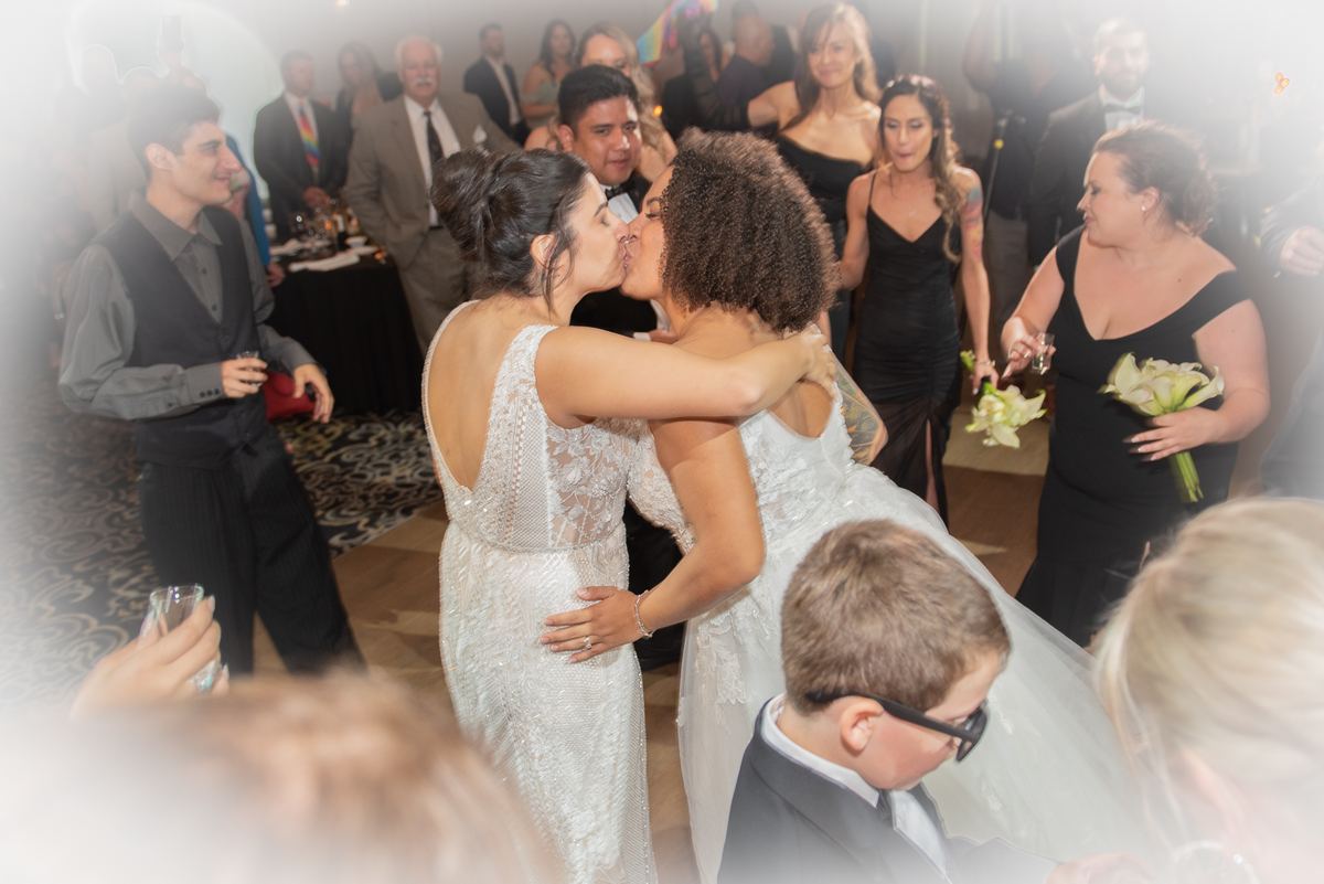 Two brides kiss at a reception surrounded by smiling guests in formal attire, creating a joyful atmosphere.