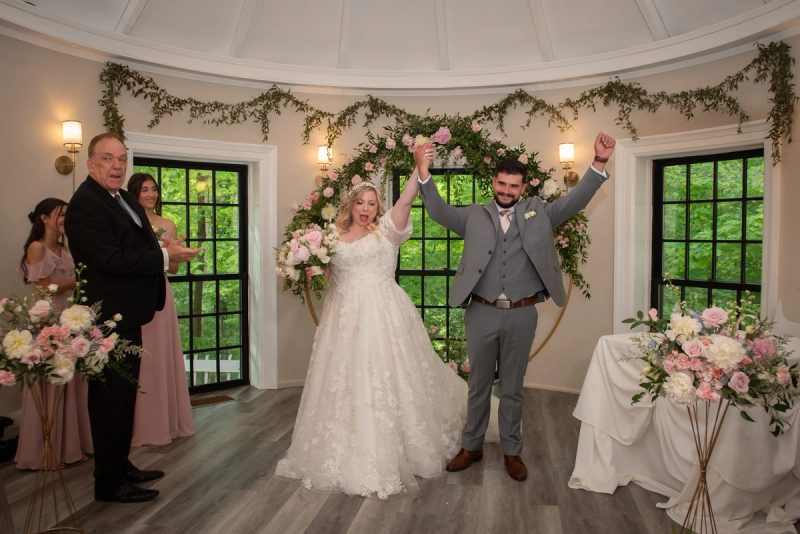 The bride and groom raise their arms joyfully in front of floral arches, surrounded by smiling guests.