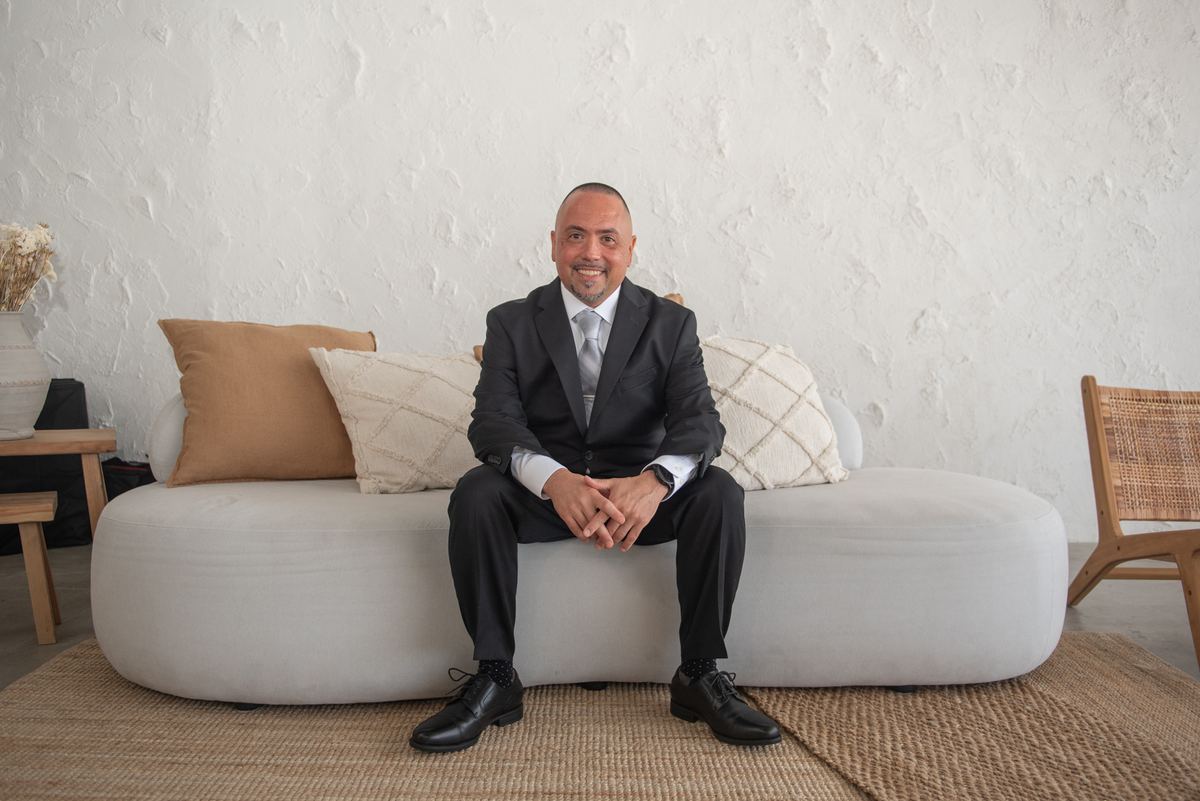 A man in a black suit smiles at the camera from a modern sofa in a minimalist room with woven pillows and jute rug.