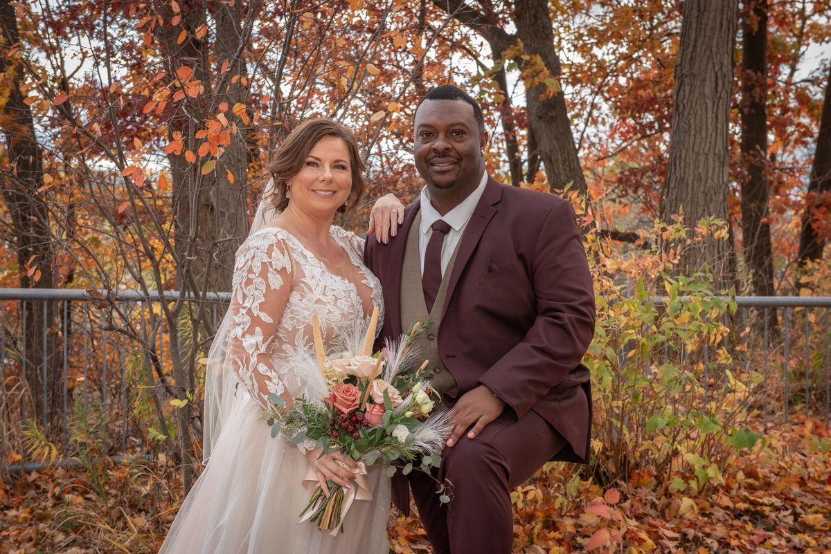 A bride in a lace gown and a groom in a burgundy suit smile amidst autumn foliage, bathed in soft daylight.