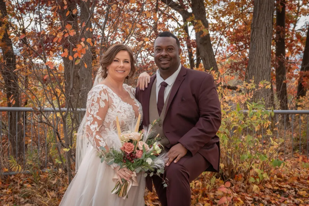 A bride in a lace gown and a groom in a burgundy suit smile amidst autumn foliage, bathed in soft daylight.