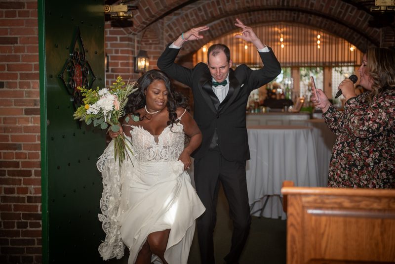 A bride and groom dance joyfully in a warmly lit indoor space with exposed brick, as a woman records the moment.