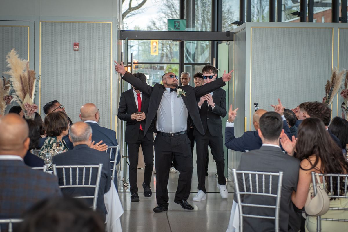 A groom joyfully strides down the aisle with outstretched arms, flanked by excited groomsmen and guests.
