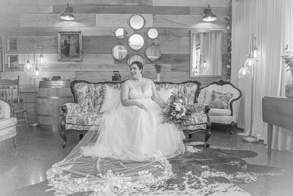 A smiling bride in a wedding gown sits on an ornate floral sofa surrounded by vintage decor and soft lighting.