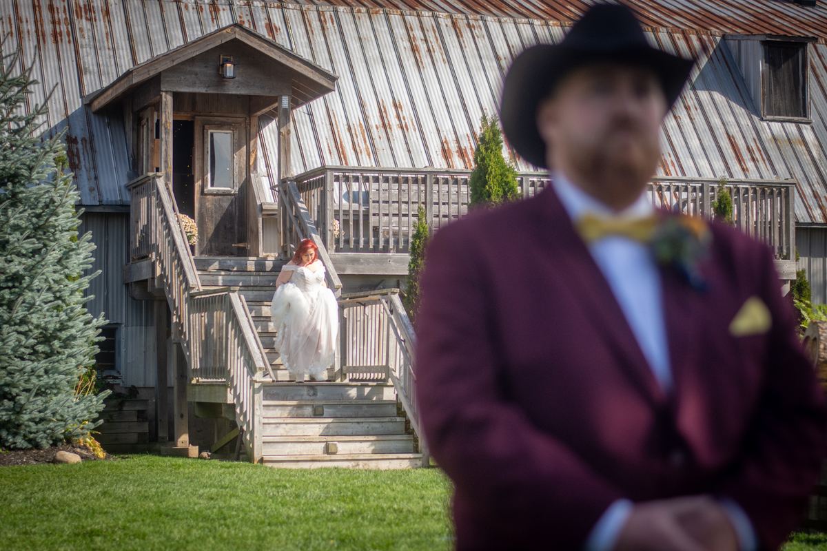 Red-haired bride in white gown descends wooden stairs toward groom in burgundy suit, set against rustic barn and greenery.