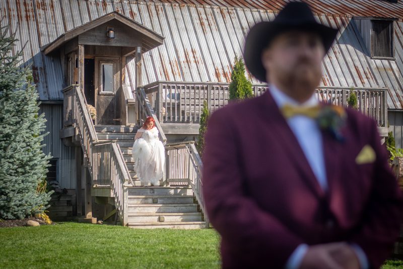 Red-haired bride in white gown descends wooden stairs toward groom in burgundy suit, set against rustic barn and greenery.