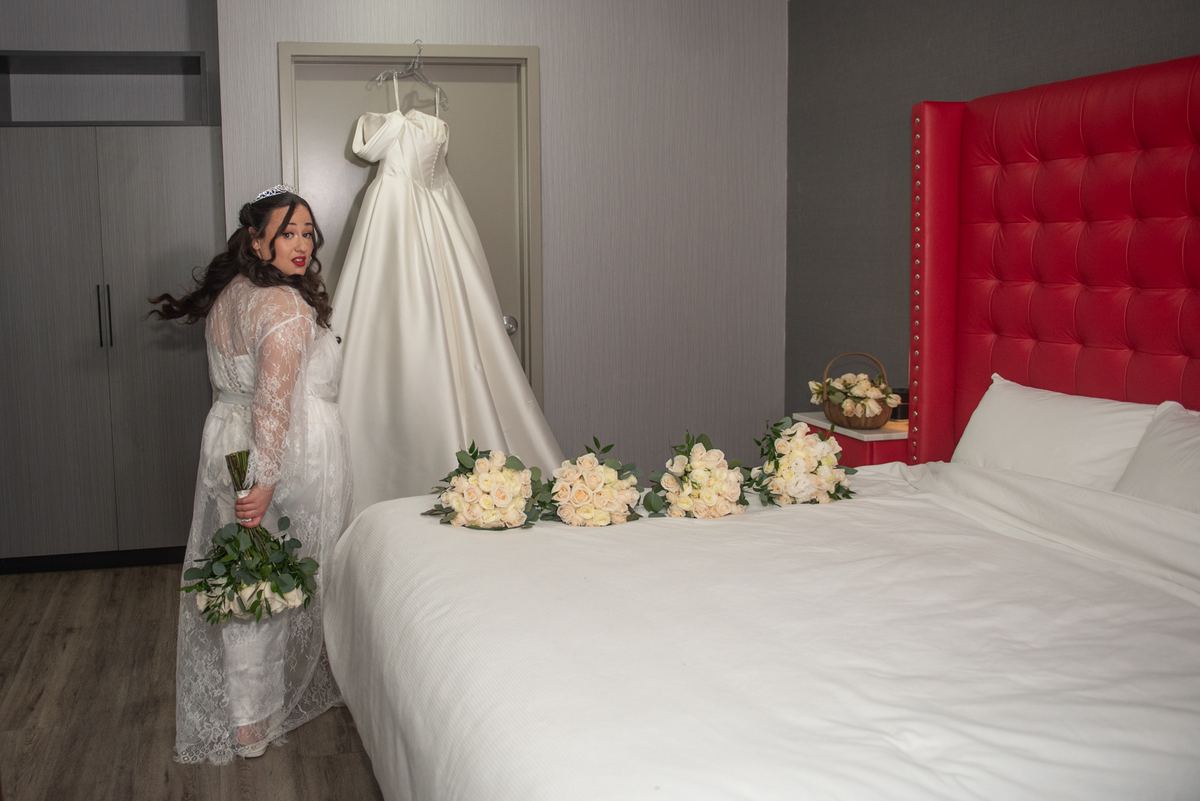 A bride stands beside a bed, holding a bouquet with her wedding gown hanging on the door and more bouquets on the bed.
