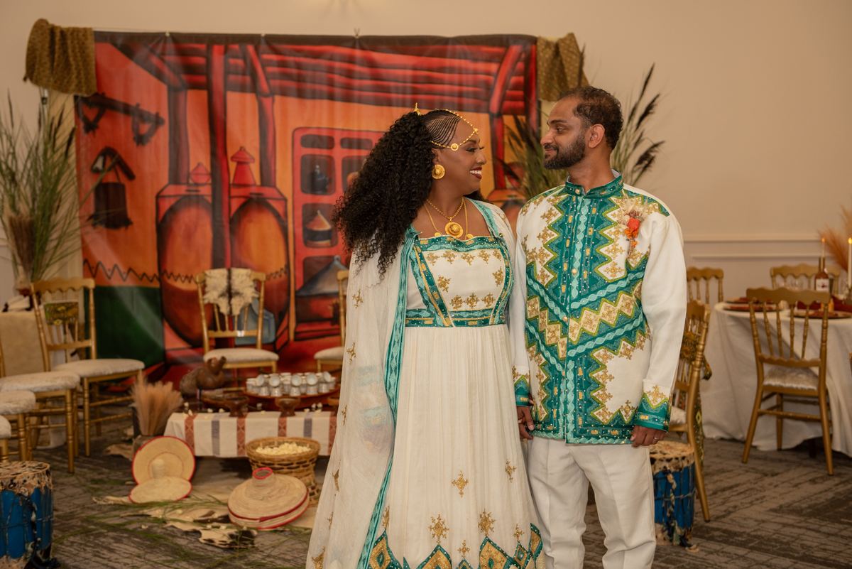 A couple in ornate gold and teal attire stands with affection in a space decorated with African architecture.