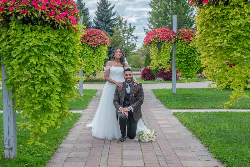 A smiling bride and kneeling groom pose outdoors amid lush greenery and pink flowers, bathed in soft daylight.