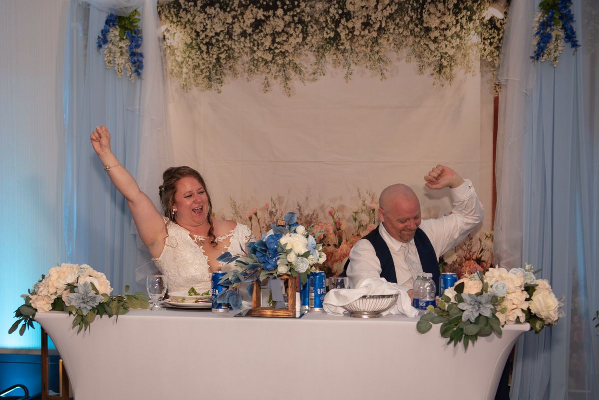 Bride and groom raise fists in celebration at reception table under floral canopy, illuminated by blue lights.